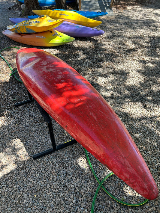 A used Perception Super Sport kayak from 4Corners Riversports is resting upside down on a stand, with several other colorful kayaks lying on the gravel in the background.