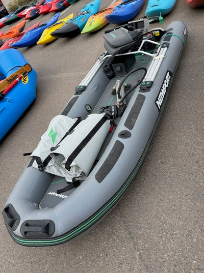 A gray Newport Demo NS130 Skiff inflatable boat is shown with a rolled-up bag, oars, and motor mount—ideal for the Newport NT300 Electric Motor. Displayed on pavement among other colorful kayaks and watercraft.