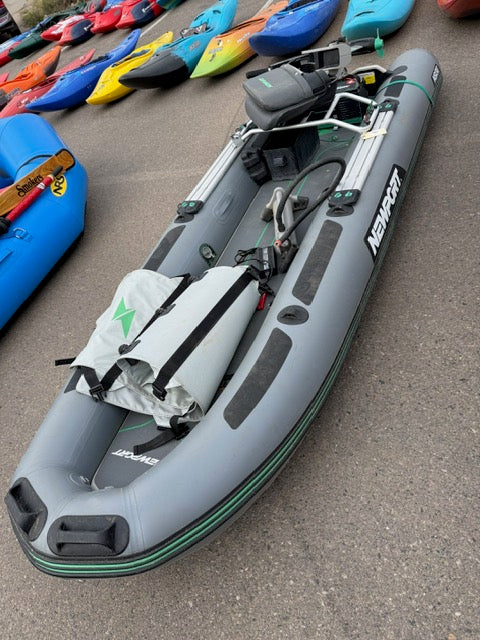 A gray Newport Demo NS130 Skiff inflatable boat is shown with a rolled-up bag, oars, and motor mount—ideal for the Newport NT300 Electric Motor. Displayed on pavement among other colorful kayaks and watercraft.