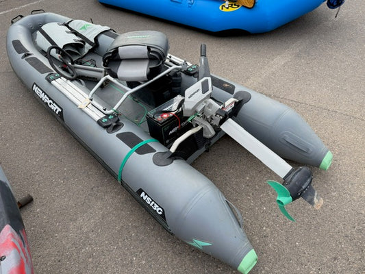 A gray Demo NS130 Skiff by Newport, equipped with a 36V lithium battery, NT300 Electric Motor, and seat, rests on asphalt beside other boats.