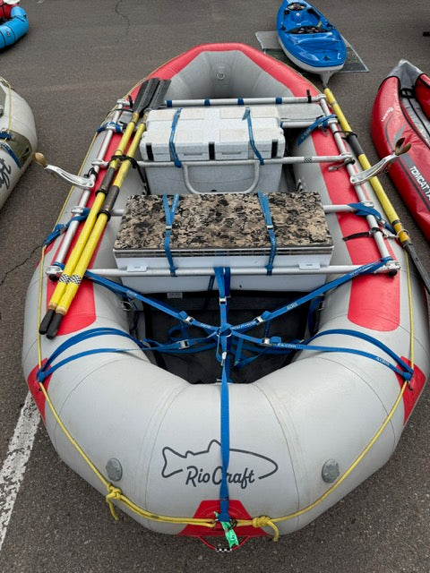 The Demo Rio Craft 14' Package inflatable raft by Rio Craft, featuring red accents, gear boxes, coolers, blue straps, and multiple oars from the expedition package, is parked on asphalt among other boats.