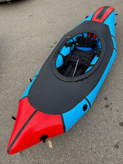 The Demo Mage LG by Alpacka is a blue and red inflatable kayak with a black seat and gear inside, featuring a Whitewater Deck, shown on a paved surface.