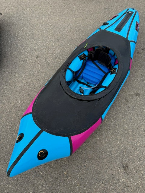 A blue and black Alpacka Demo Valkyrie LG kayak with pink accents sits on asphalt, revealing its cockpit and interior—a great choice for those seeking a quality used packraft from a trusted demo fleet.