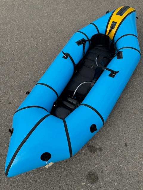 A used demo Alpacka Demo Gnarwhal XL inflatable kayak, blue and black with a yellow front section, is placed on a paved surface.