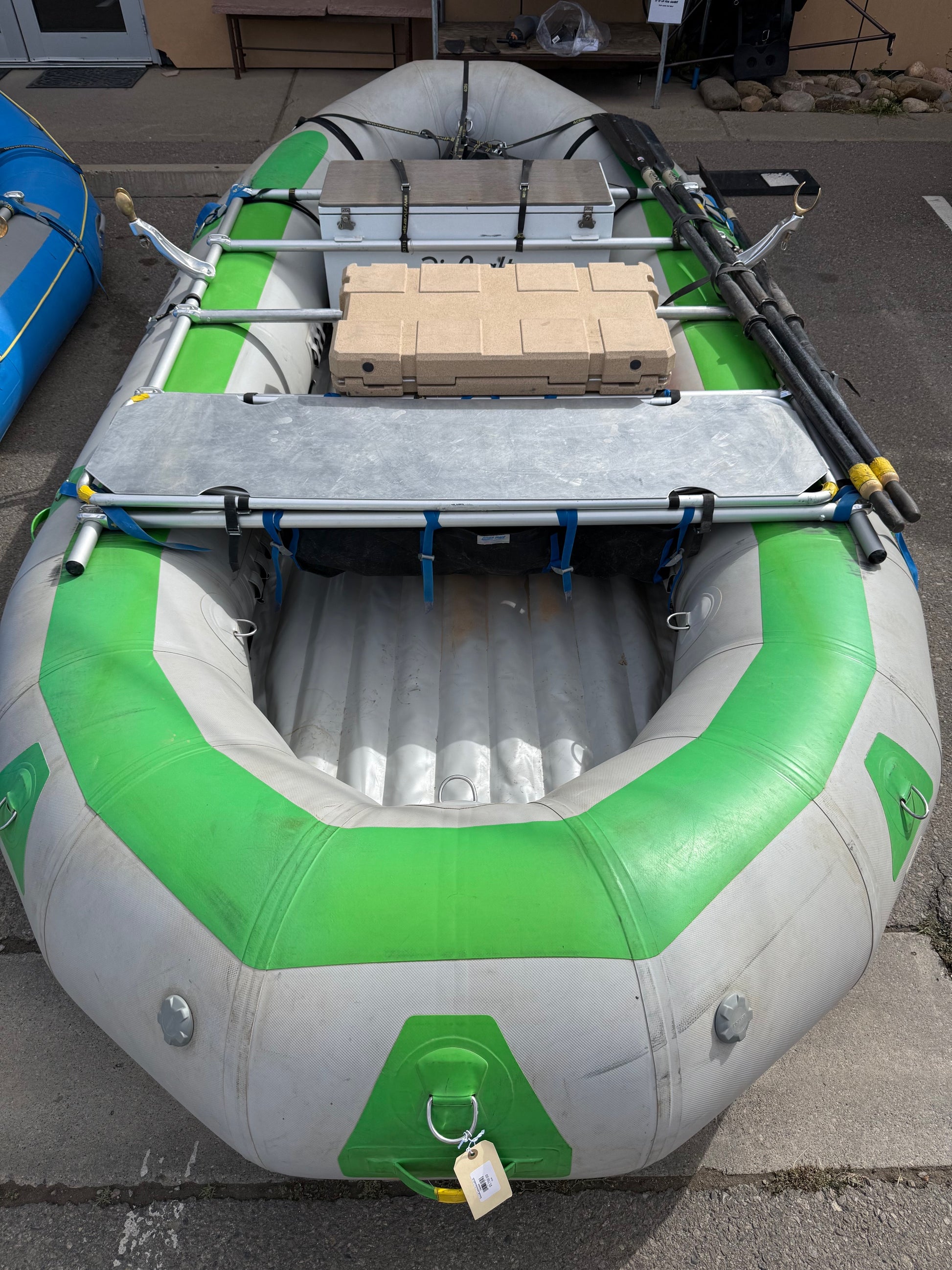 A Demo Rio Craft 16' Package raft by Rio Craft in gray and green, equipped with oars and stacked tan bricks on an NRS Frame, is set on a paved surface.