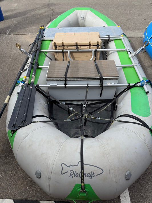A Demo Rio Craft 16' Package by Rio Craft—featuring a gray and green raft with NRS Frame, two black oars, gear, and box—sits on a paved surface.