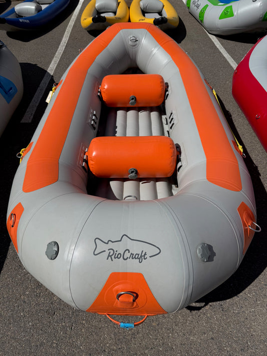A gray and orange Rio Craft 10.5' Demo inflatable raft with two orange seats is shown on asphalt among other rafts.