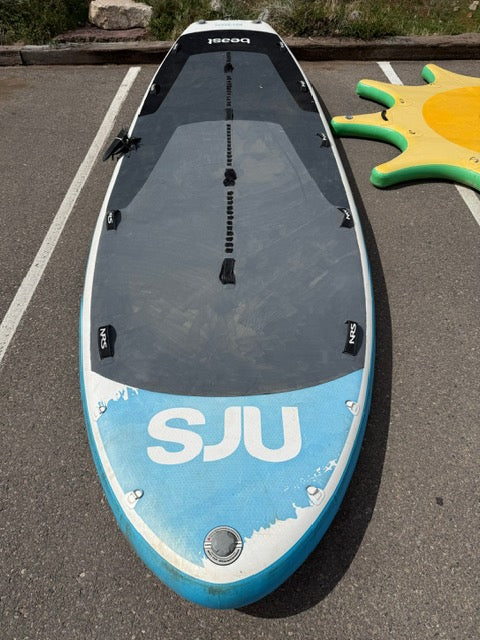 A used NRS Demo Beast SUP with "SJU" on the front rests on the ground beside a yellow and green inflatable in a parking lot.