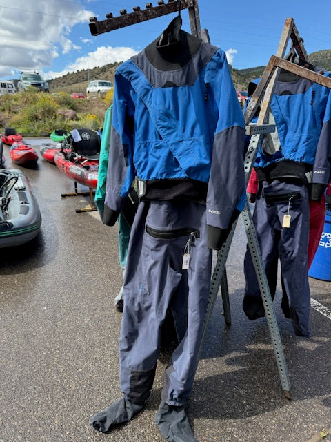 A Demo Drysuit - NRS Axiom Men's SM by NRS hangs on a metal rack outdoors, with kayaks and inflatable boats in the background.