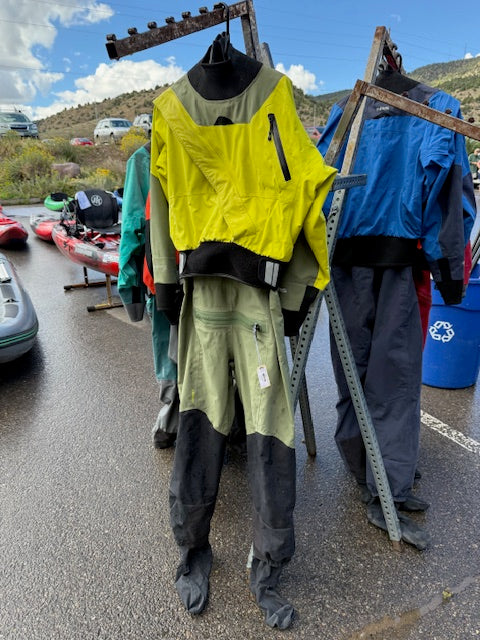 A yellow and green NRS Axiom Men's LG Demo Drysuit by NRS hangs on a metal rack outdoors, with kayaks, other demo drysuits, and mountains visible in the background.