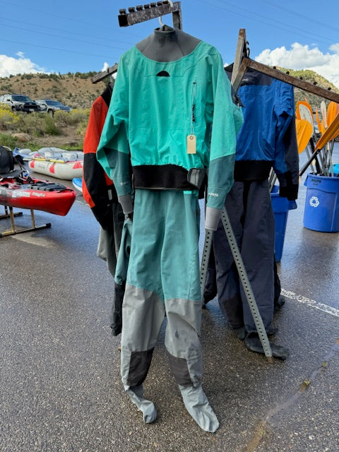 A green and gray NRS Phenom Women's XL drysuit hangs on a metal rack outside, with other drysuits, kayaks, and outdoor gear available for rental in the background.