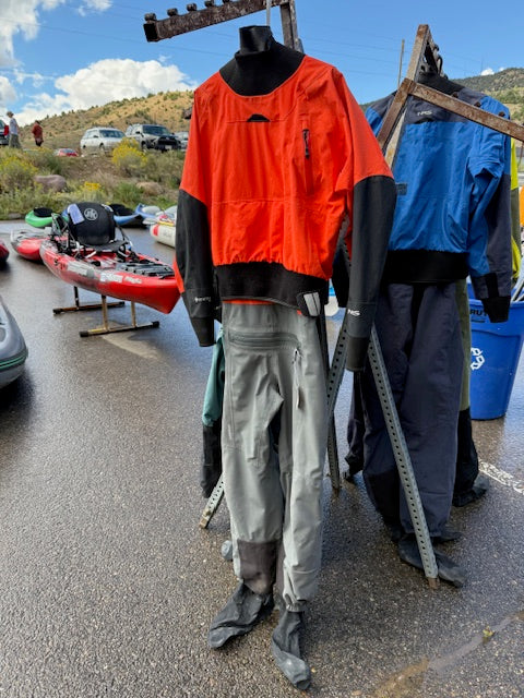 An NRS Phenom Men's LG Demo Drysuit by NRS is displayed on a mannequin at an outdoor event, with kayaks and vehicles visible in the background.