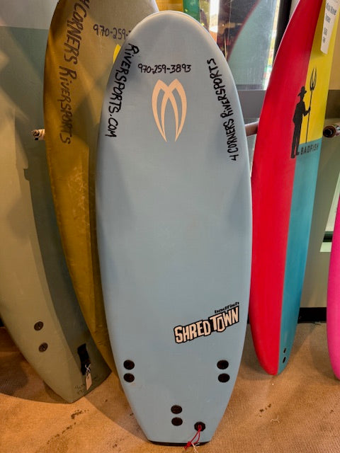 A light blue Badfish Demo Shredtown bodyboard with black fin holes and text stands upright among colorful boards in a shop—ideal for those seeking a reliable soft top river surf board.