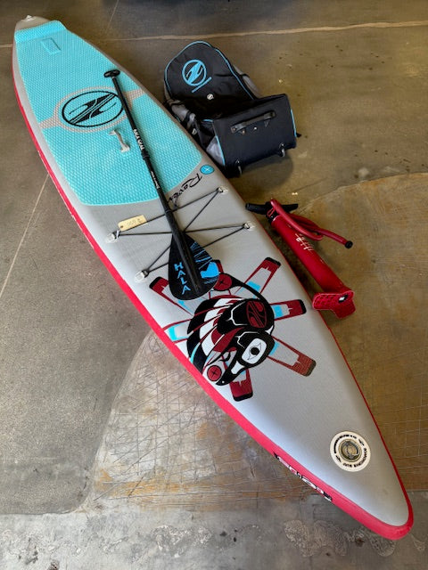 The Boardworks Used Raven 12'6 Inflatable Paddle Board in blue and red comes with a carbon paddle, air pump, repair kit, and carrying bag—shown organized on concrete. Ideal for aquatic adventures.
