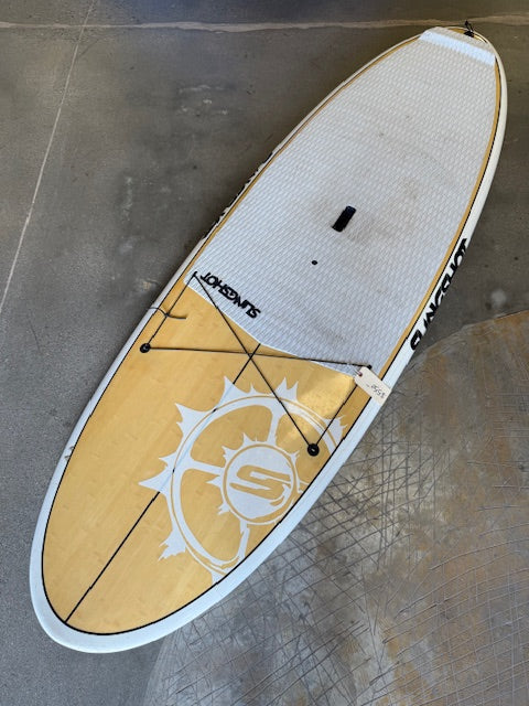 The 4Corners Riversports Consignment Slingshot Space Pickle SUP, showcasing a white and wood-grain finish with a "SUNSATION" logo, black handle, and deck bungees, is displayed elegantly on a concrete floor.