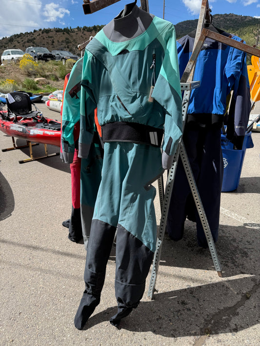 A green and black NRS Axiom Women's LG demo drysuit hangs on a metal rack outdoors, with kayaks and other rental drysuits visible in the background on a sunny day.