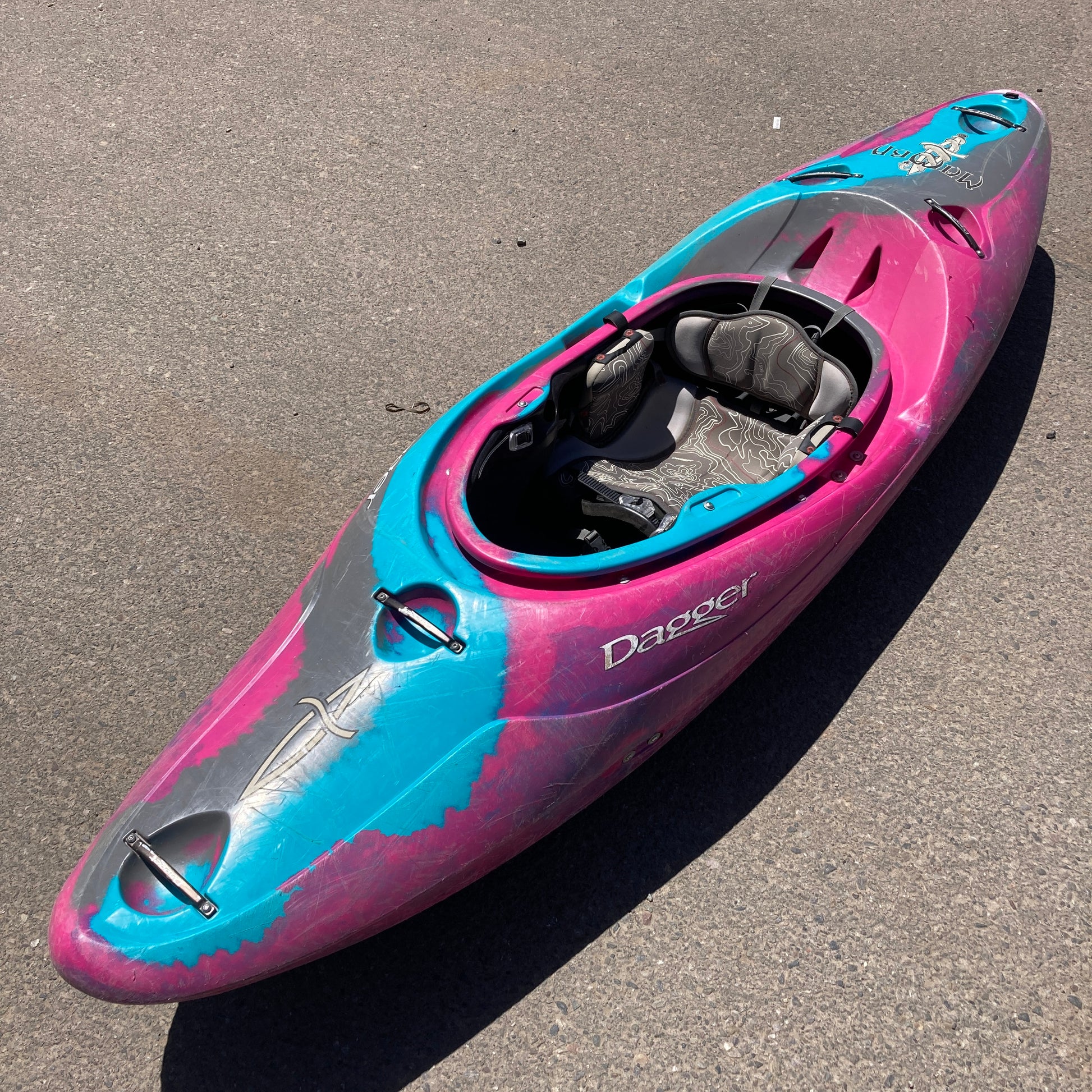 A vibrant pink and blue Dagger Mamba 7.6 kayak with the brand "Dagger Mamba 7.6" on its side, resting on an asphalt surface in sunlight.