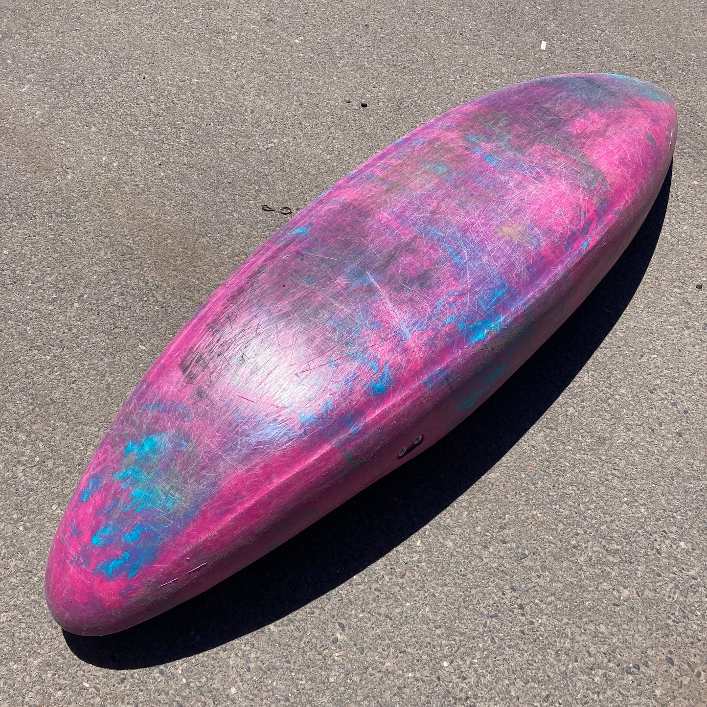 A colorful, well-worn Consignment Dagger Mamba 7.6 kayak with pink and blue patterns lying on sunlit asphalt.