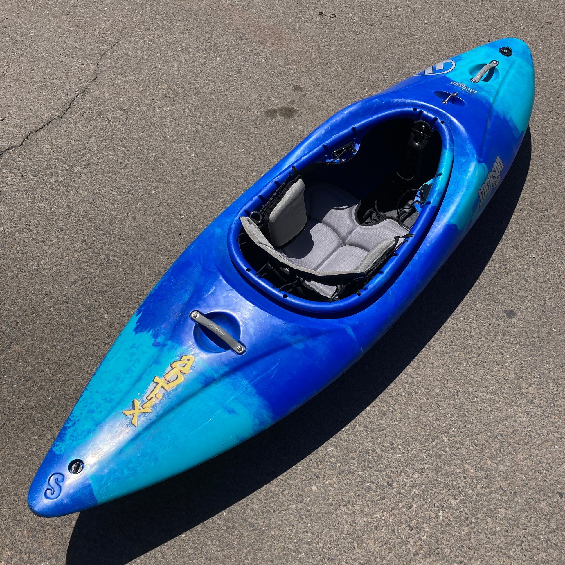 A blue Consignment Jackson Antix S whitewater kayak by Jackson Kayak with visible river rash and a brand logo, positioned on a sunlit asphalt surface.