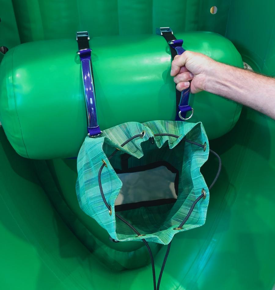 A hand grips a strap connected to the Thwart Bag by Down River Equipment, a green inflatable structure with a UV-resistant, multi-mesh fabric bag.