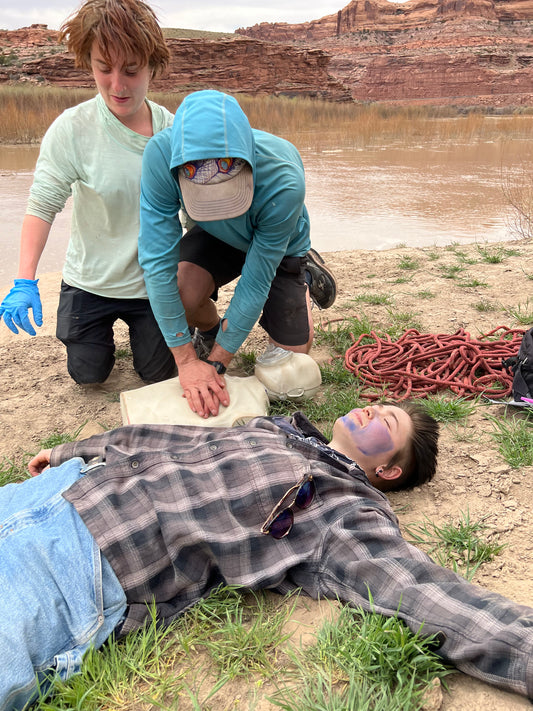 Two people with CPR & AED Training from 4CRS Paddle School perform CPR on a person by a riverbank, with climbing rope and a resuscitation bag nearby.