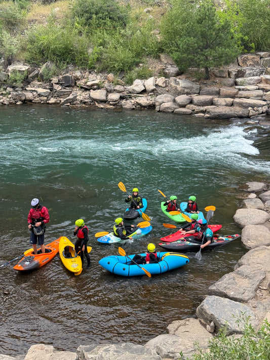 A group in colorful kayaks and safety gear gathers by a rocky riverbank, getting ready for or wrapping up the Send-It Summer Finale by 4CRS Paddle School.
