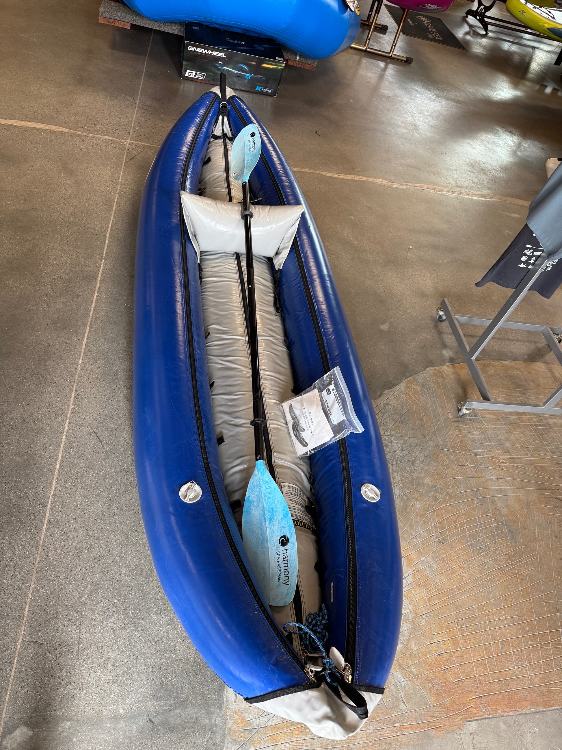 The Consignment Tributary Tomcat Solo by 4CRS, a blue inflatable kayak known as a ducky, rests gracefully on the concrete floor. Designed for solo adventurers with Harmony Paddles, it promises an exciting ride for those eager to explore waterways.