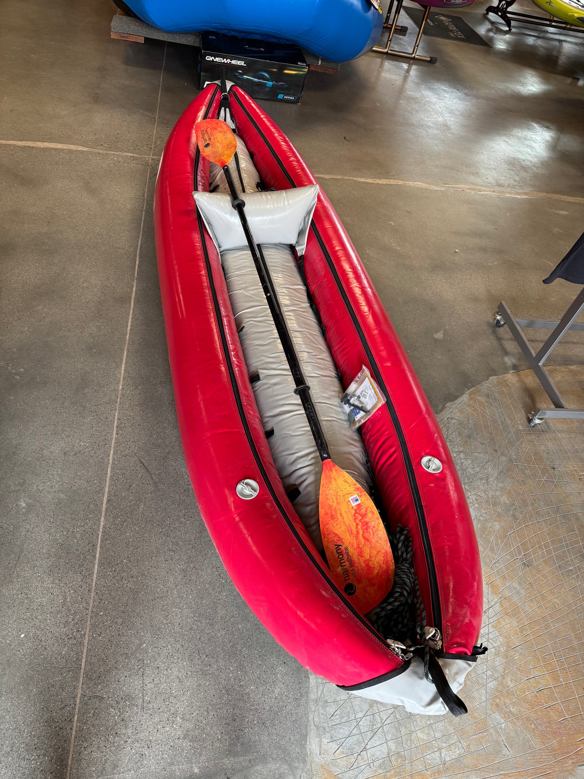 The 4CRS Consignment Tributary Tomcat Solo, a red inflatable kayak with a gray seat, is displayed indoors. Two orange paddles lay across its center, emphasizing its adventure readiness.