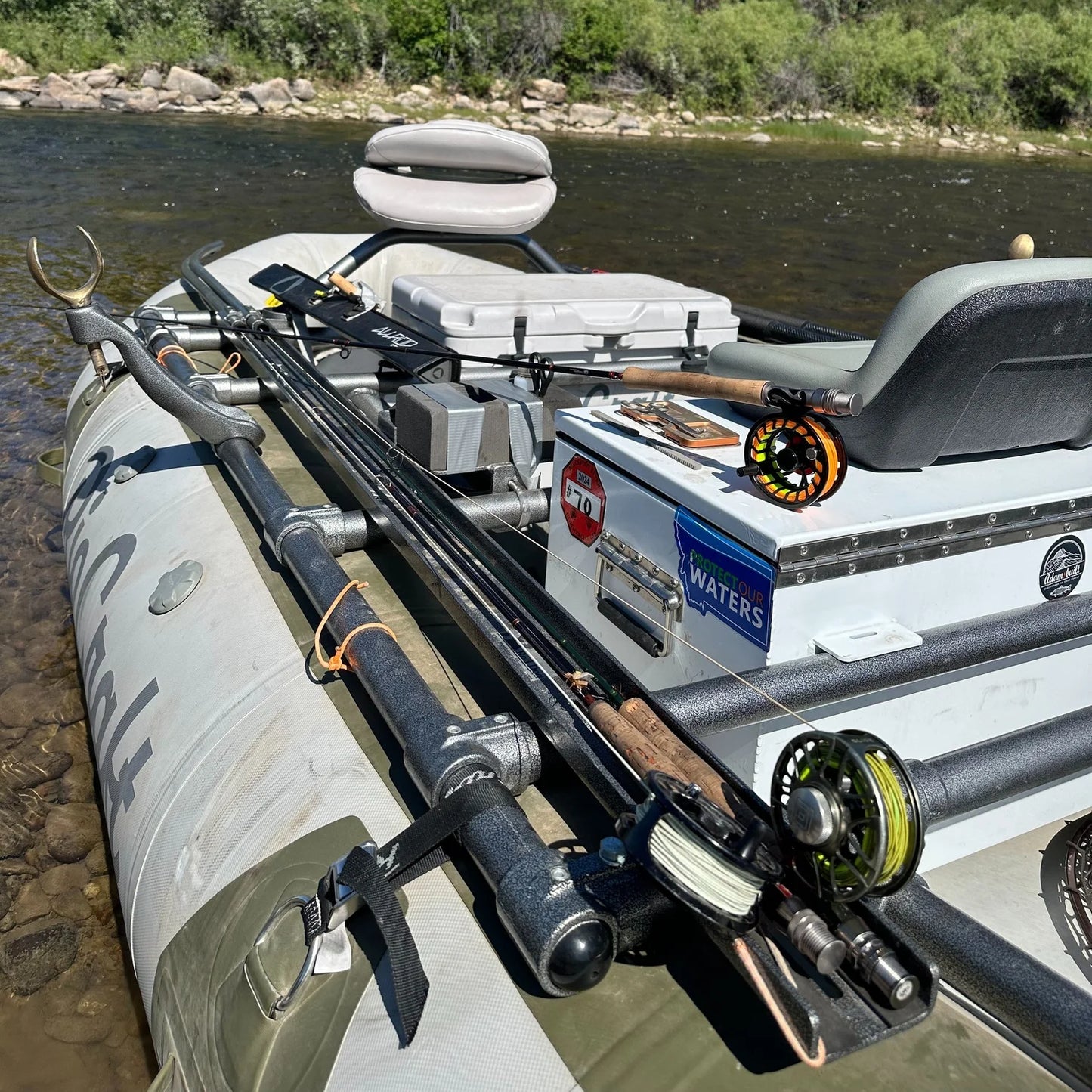 A Rio Craft raft equipped with a mounted seat, reels, cooler, gear box, and the Rod Dog Rod Holder floats on a shallow river near a grassy, wooded bank.