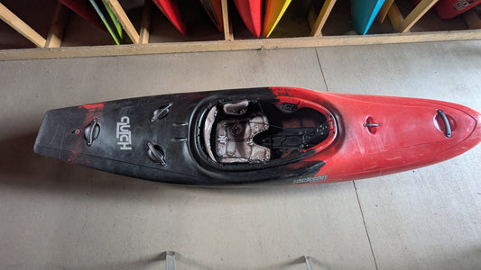 A red and black Jackson Kayak Demo Clutch 2025 sits on a concrete floor, labeled with the brand’s name, while other colorful demo kayaks are stored vertically in the background.
