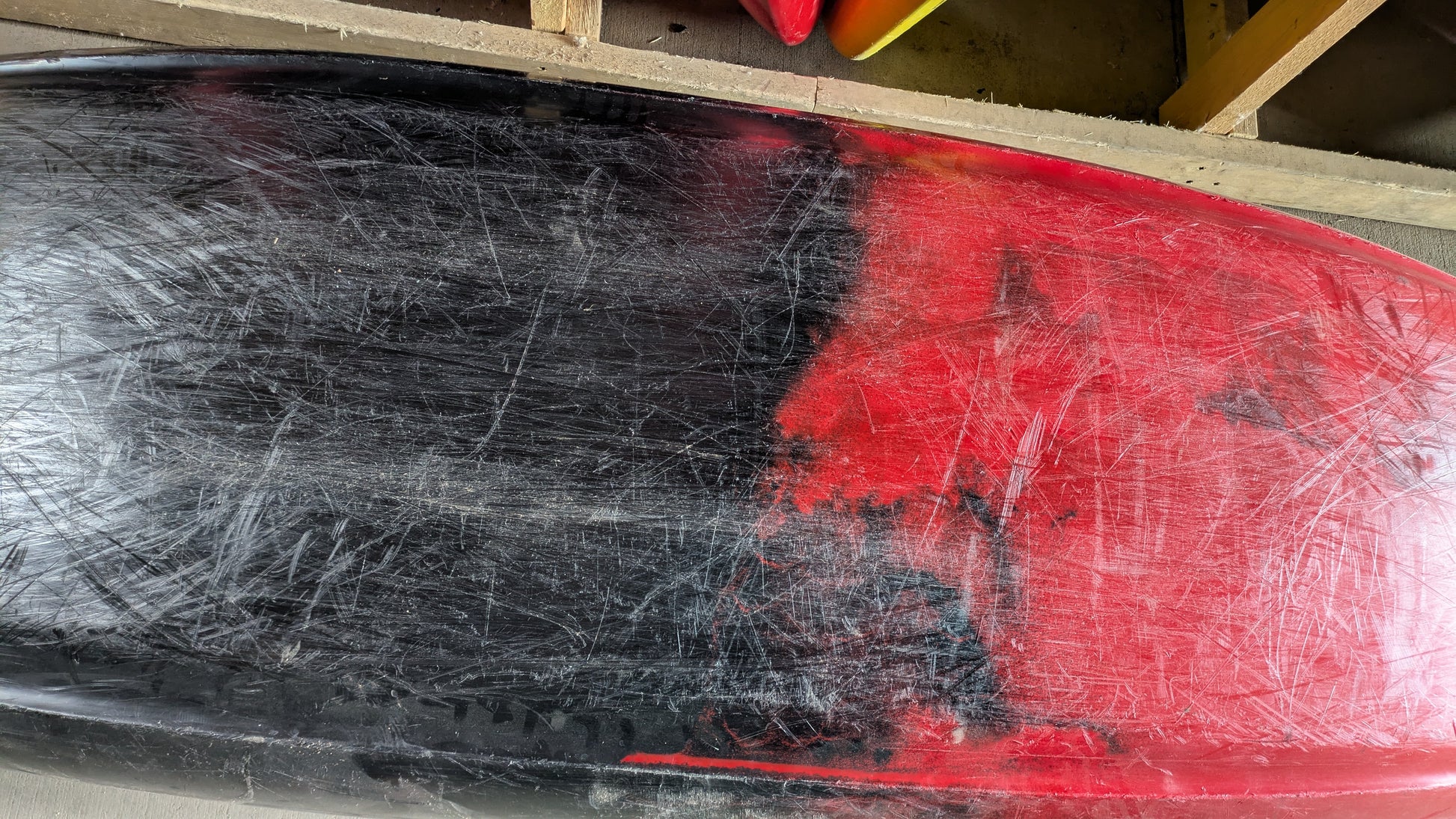 Close-up of a used Jackson Kayak Demo Clutch 2025 hull with heavy black and red scratches beneath a wooden rack.