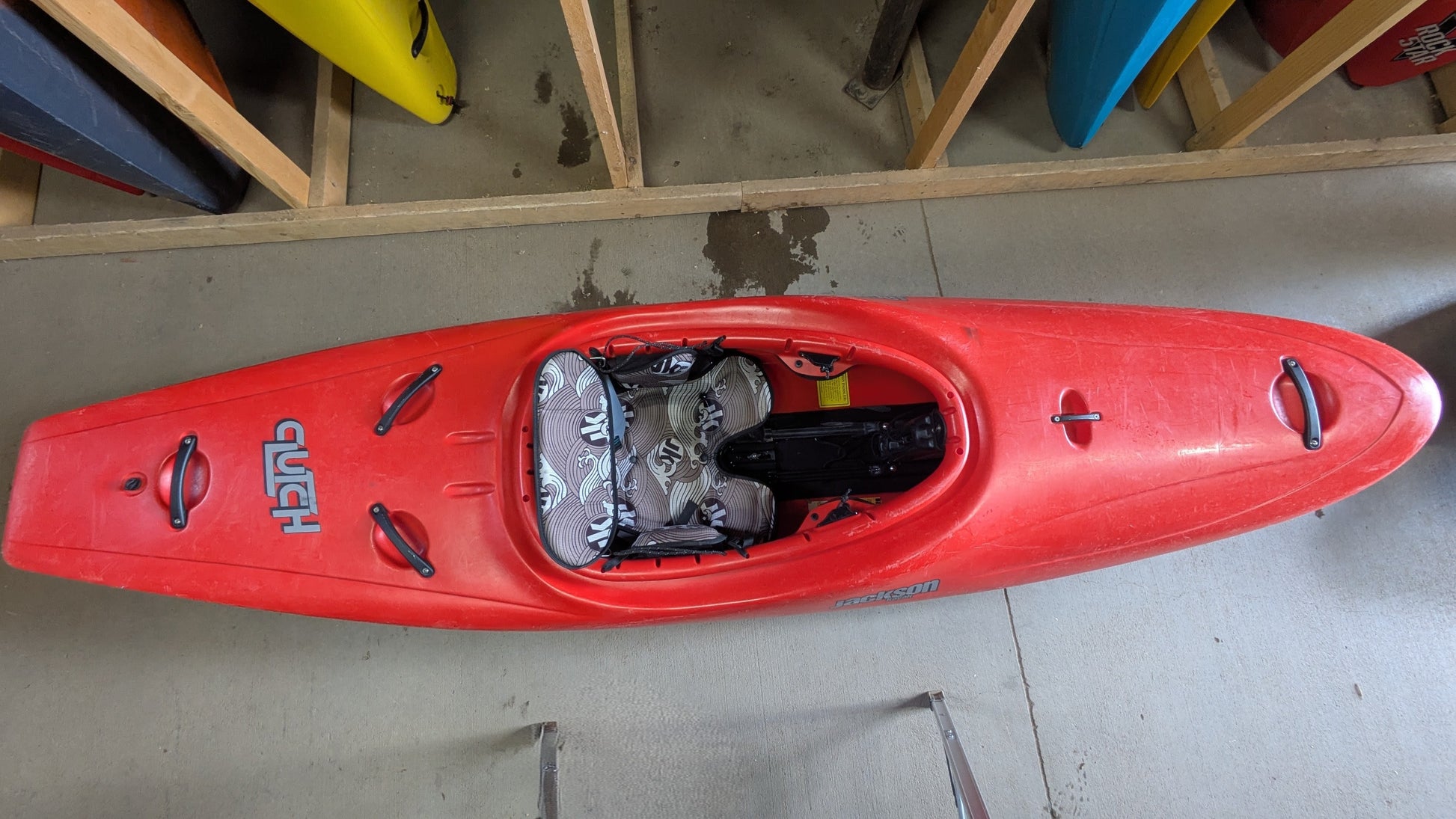 A red Jackson Kayak Demo Clutch 2025 is stored indoors on a concrete floor among other kayaks on racks, with its cockpit and seat area visible from above.