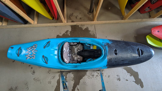 A Jackson Kayak Demo Gnarvana MD in blue, labeled "Gnarvana," rests on a concrete floor amid other demo fleet kayaks stored vertically on racks.