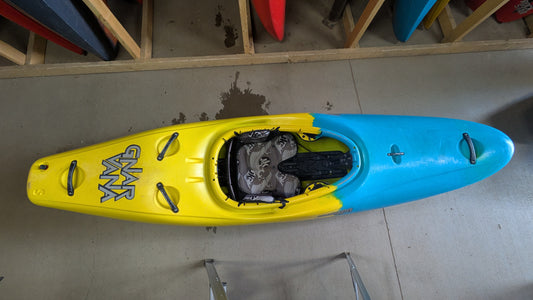 A Jackson Kayak Demo Gnarvana SM in blue and yellow from our demo fleet rests on a concrete floor, with shelves of other kayaks nearby. The interior and seat of this used kayak are clearly visible from above.