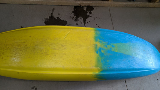 A yellow and blue Demo Gnarvana SM by Jackson Kayak, from a demo fleet, lies upside down on a gray floor with dark stains in the background.
