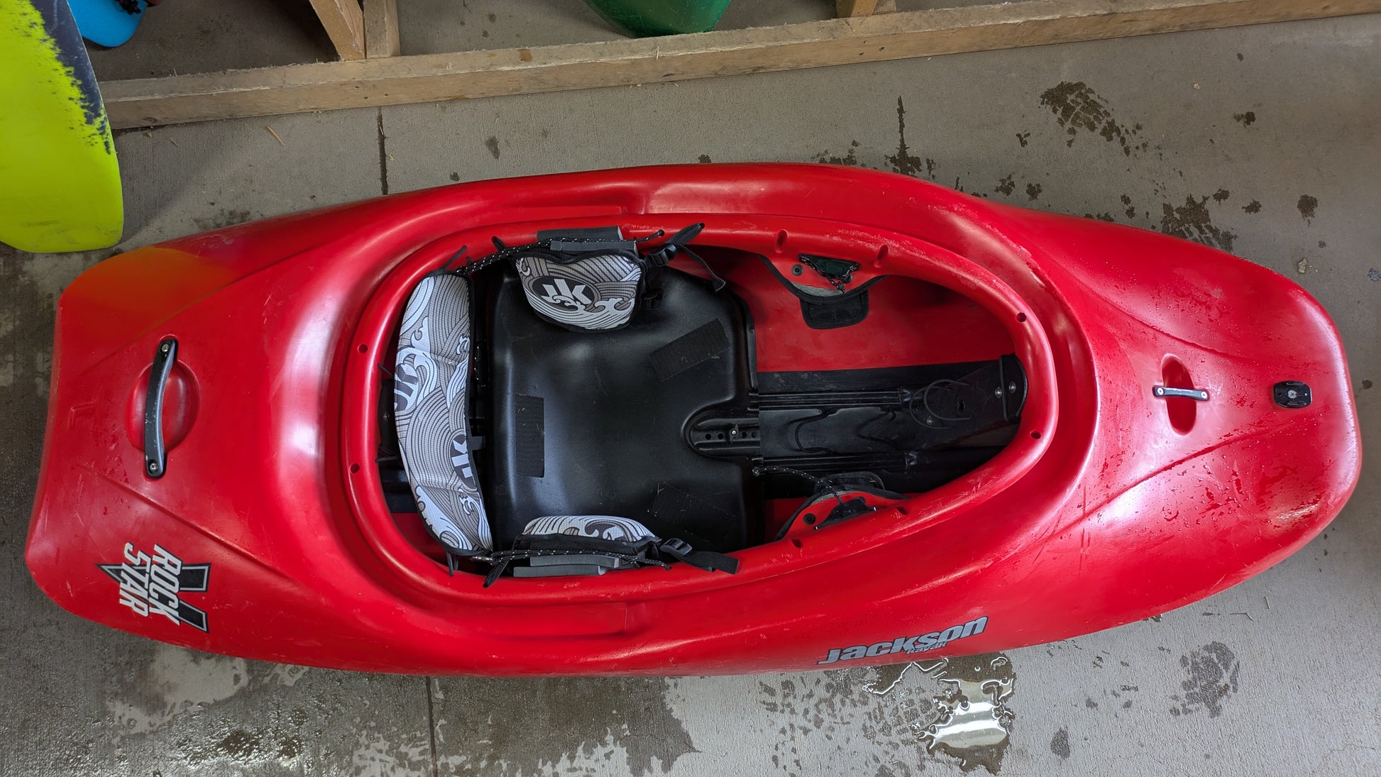 A red used Jackson Kayak Demo Rockstar V LG is upright on a wet concrete floor, displaying the cockpit and seat from above.