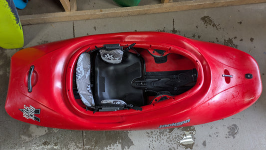 A red used Jackson Kayak Demo Rockstar V LG is upright on a wet concrete floor, displaying the cockpit and seat from above.