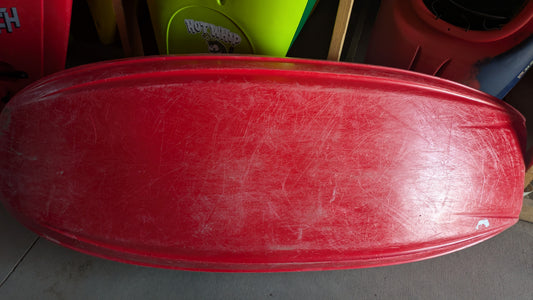 A Jackson Kayak Demo Rockstar V MD with noticeable scratches rests upside down on a concrete floor; colorful sleds, another used kayak, and various objects are partially visible in the background.