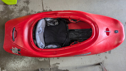 A Jackson Kayak Demo Rockstar V SM in red rests on a concrete surface, seen from above with its seat, footrests, and cockpit interior visible.