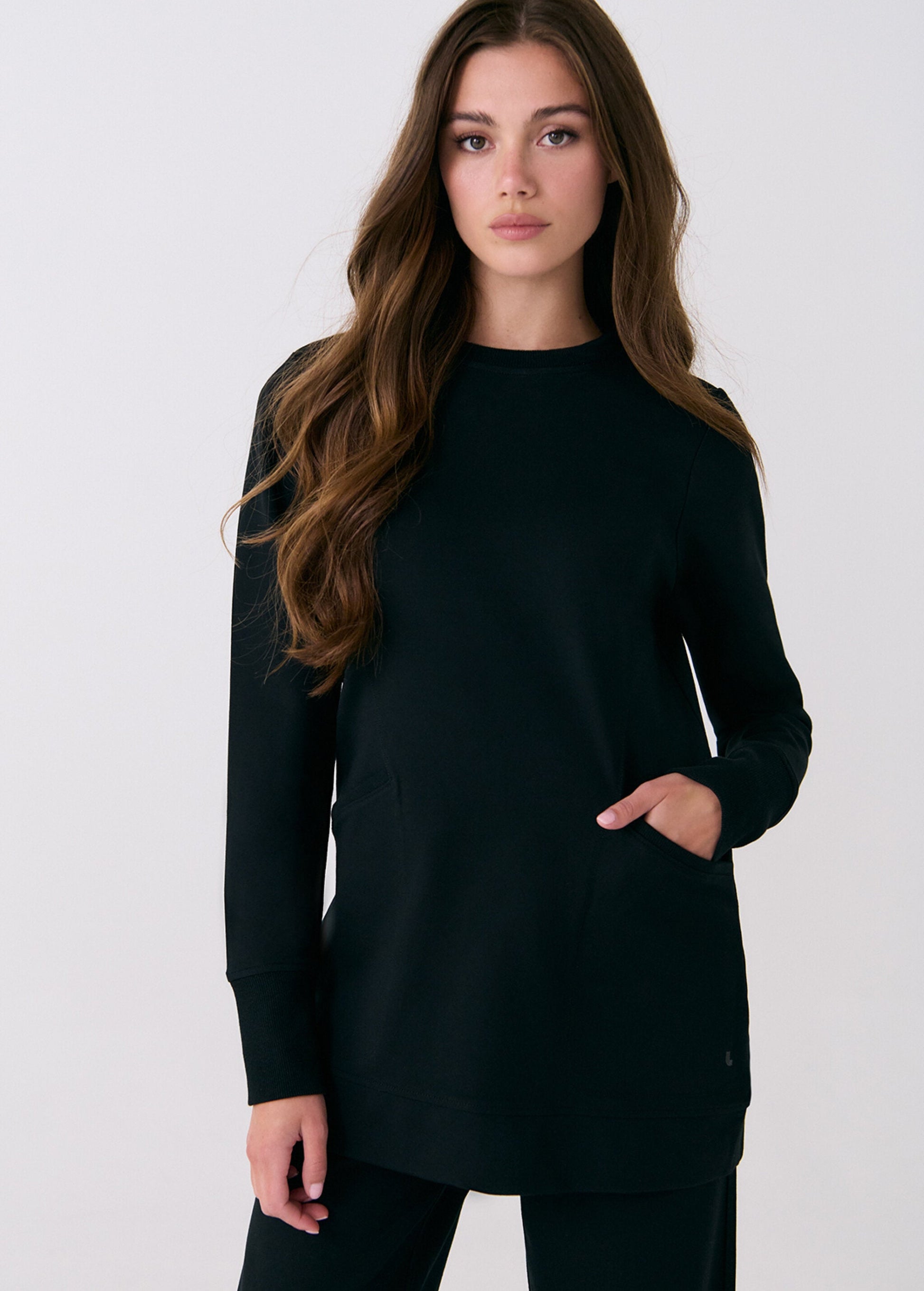 A woman with long brown hair wears the Lole Mindset Tunic in black, featuring a classic fit, front pockets, and long sleeves, paired with black pants against a plain white background.