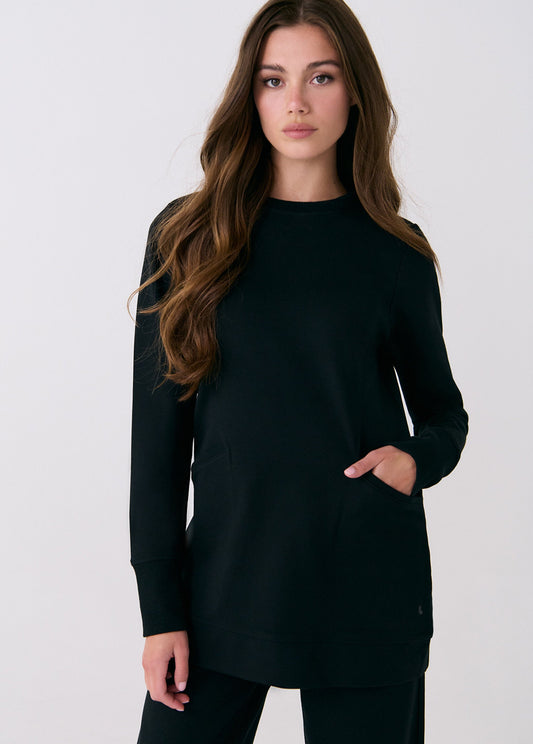 A woman with long brown hair wears the Lole Mindset Tunic in black, featuring a classic fit, front pockets, and long sleeves, paired with black pants against a plain white background.