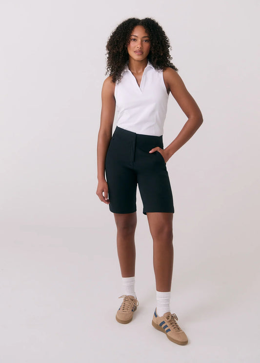 A woman stands against a plain background wearing a sleeveless white top, Lole Miles Bermuda Shorts in black with zippered pockets, white socks, and beige sneakers with blue stripes.