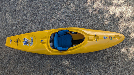 A used LiquidLogic Demo Sweet Ride kayak with yellow hull and blue seat padding is shown on a gravel surface with tree shadows. This stylish and comfortable kayak is ready for your next adventure.
