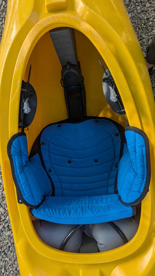 Top view of a LiquidLogic Demo Sweet Ride kayak: yellow body, blue padded seat insert, footrests, and gravel in the background.