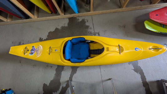 A yellow LiquidLogic Demo Powerslide kayak with a blue seat sits on a concrete floor, surrounded by other colorful kayaks stored upright on racks.