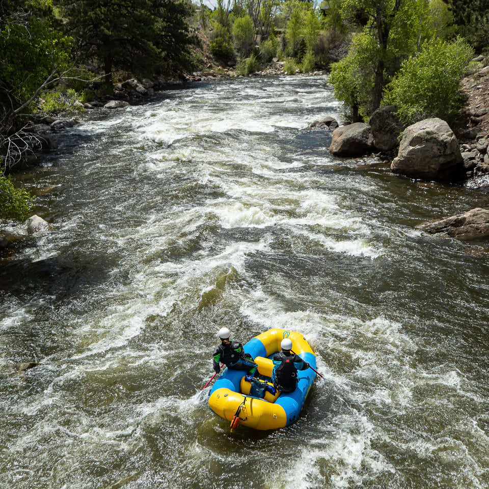 Two people in helmets paddle a Badfish Ark 10'6 raft, colored yellow and blue, down a fast, rocky river surrounded by trees and greenery.