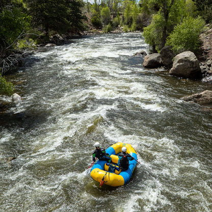 Two people in helmets paddle a Badfish Ark 10'6 raft, colored yellow and blue, down a fast, rocky river surrounded by trees and greenery.