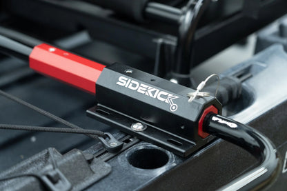 A red and black device labeled "SideKick Kayak Wheel System" is mounted on metal tubing with a lock pin. Produced by Native Watercraft, this wheel system ensures smooth kayak transport alongside your surrounding black equipment.