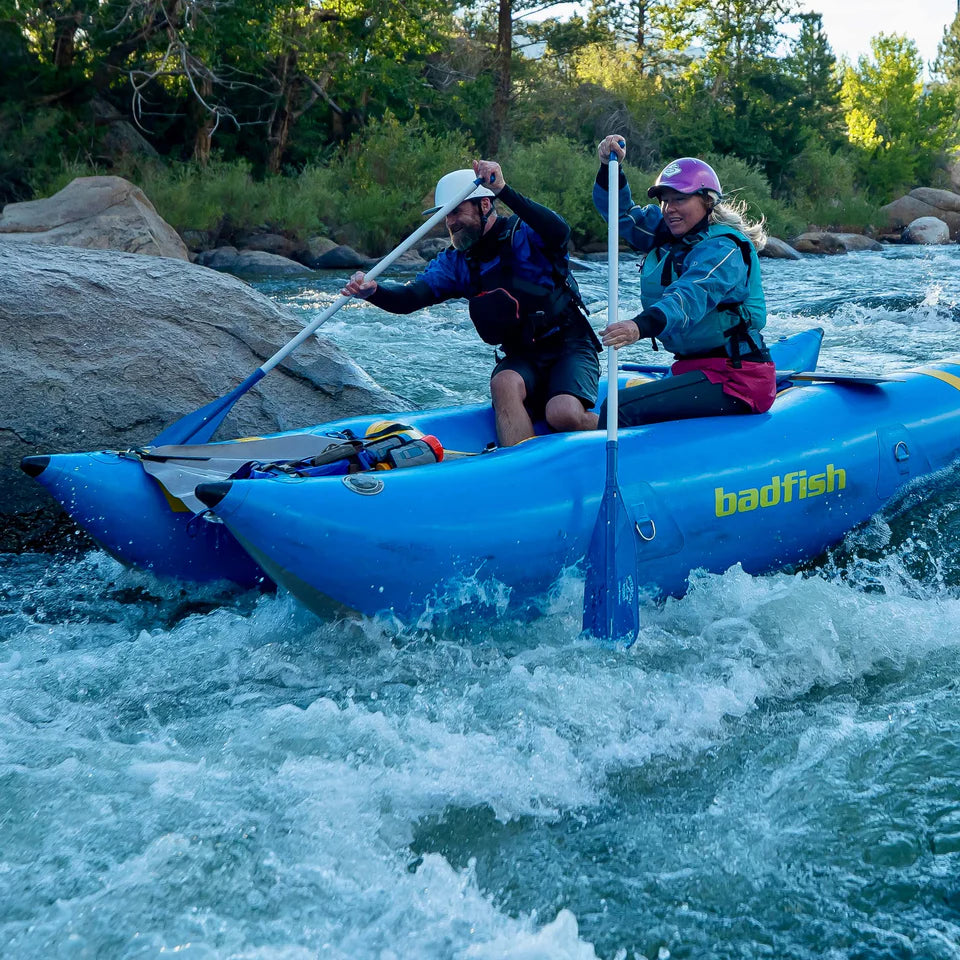 Two people in helmets and life jackets paddle a blue Badfish Hammer Cat inflatable boat through thrilling whitewater rapids. The Hammer Cat is constructed with DuraClear fabric for durability and performance.
