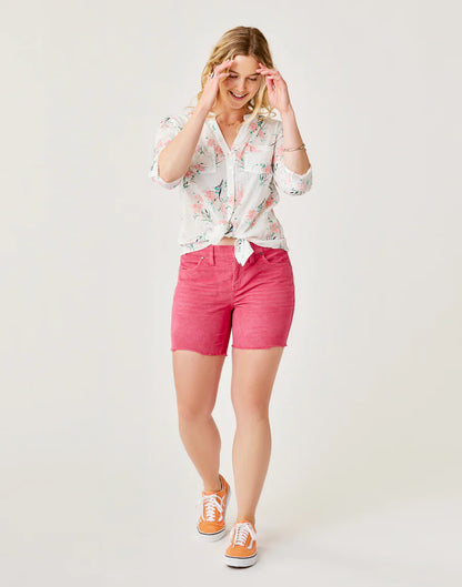 A woman smiles while walking, wearing a floral button-up shirt, Carve's Oahu 6" Short in pink stretch cotton corduroy, and orange sneakers against a plain light background—perfect for your vacation wardrobe.